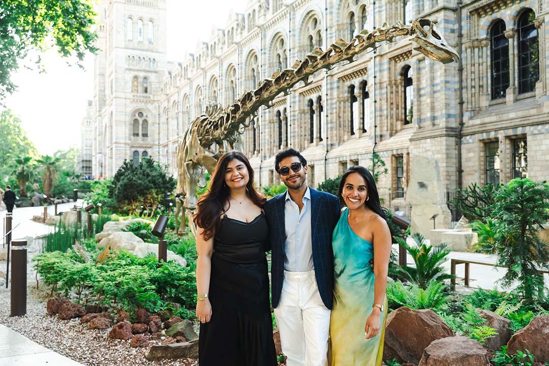 Three students smiling in front of a large grand building and a dinosaur skeleton, the woman on the left is wearing a long black dress, the man in the middle is wearing a blue blazer with white trousers, and the woman on the right is wearing a long teal and yellow gradient dress