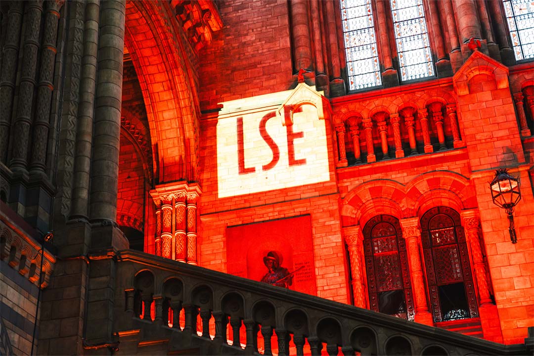 The LSE University logo projected onto the brick wall of the Natural History Museum, red lights are making the stairway glow